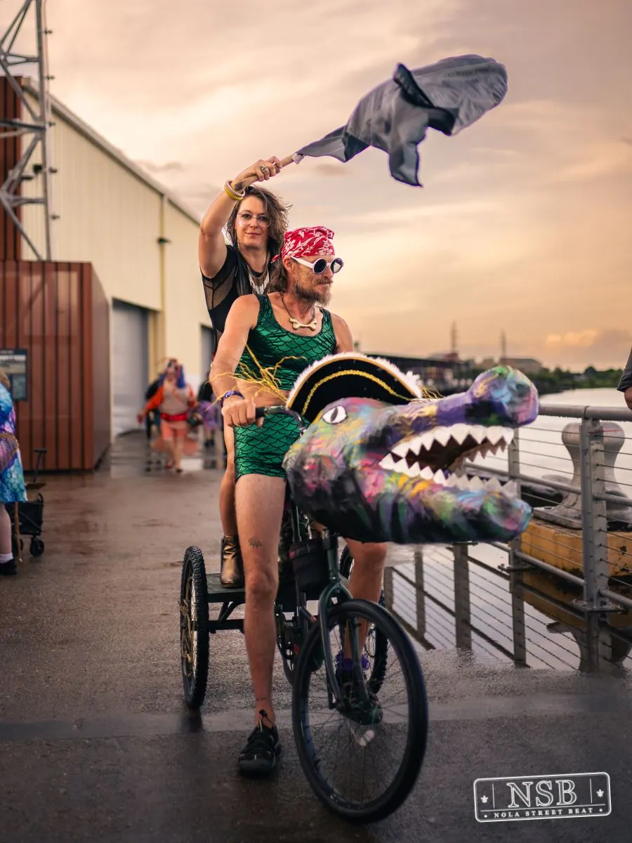 A costumed rider on a decorated sea-creature bicycle beside the Mississippi River at the 2026 New Orleans Mermaid Parade