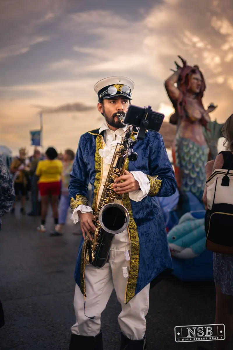 A saxophonist playing beside the river among costumed participants at the 2026 New Orleans Mermaid Parade