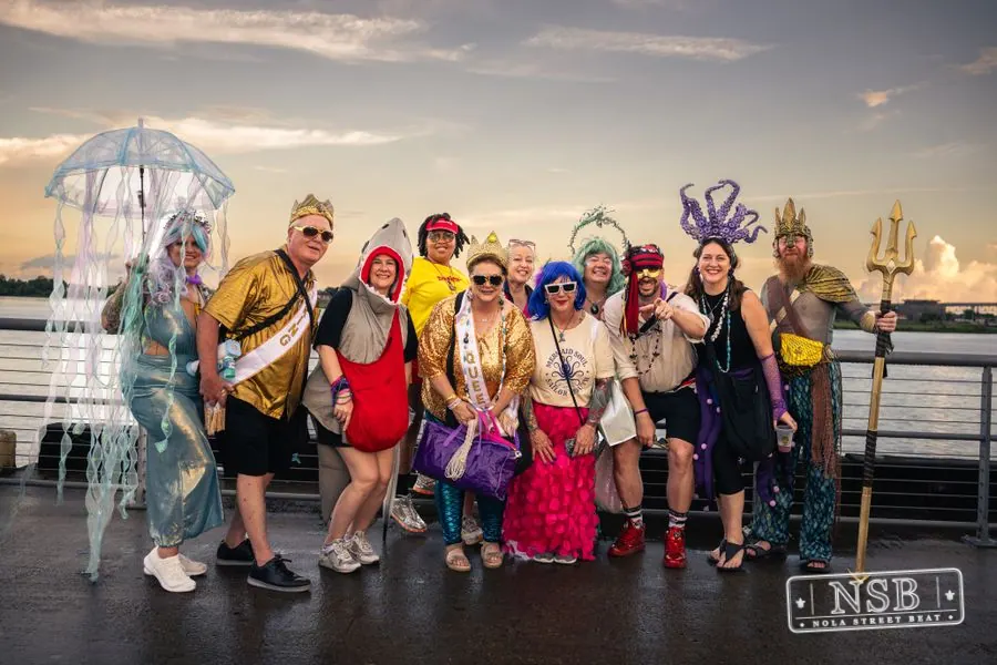A group of musicians and sea-themed parade participants beside the river at the 2026 New Orleans Mermaid Parade