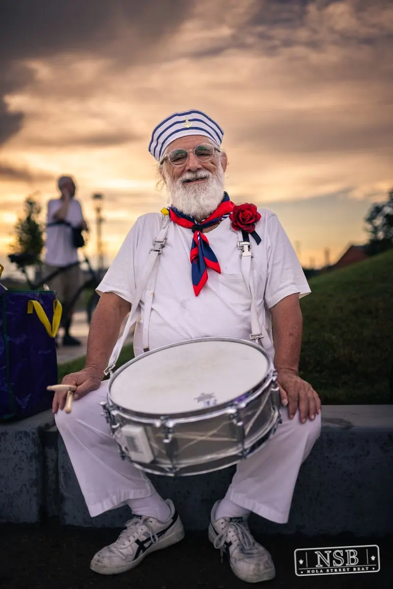 A smiling drummer wearing a white shirt and red neckerchief at the 2026 New Orleans Mermaid Parade