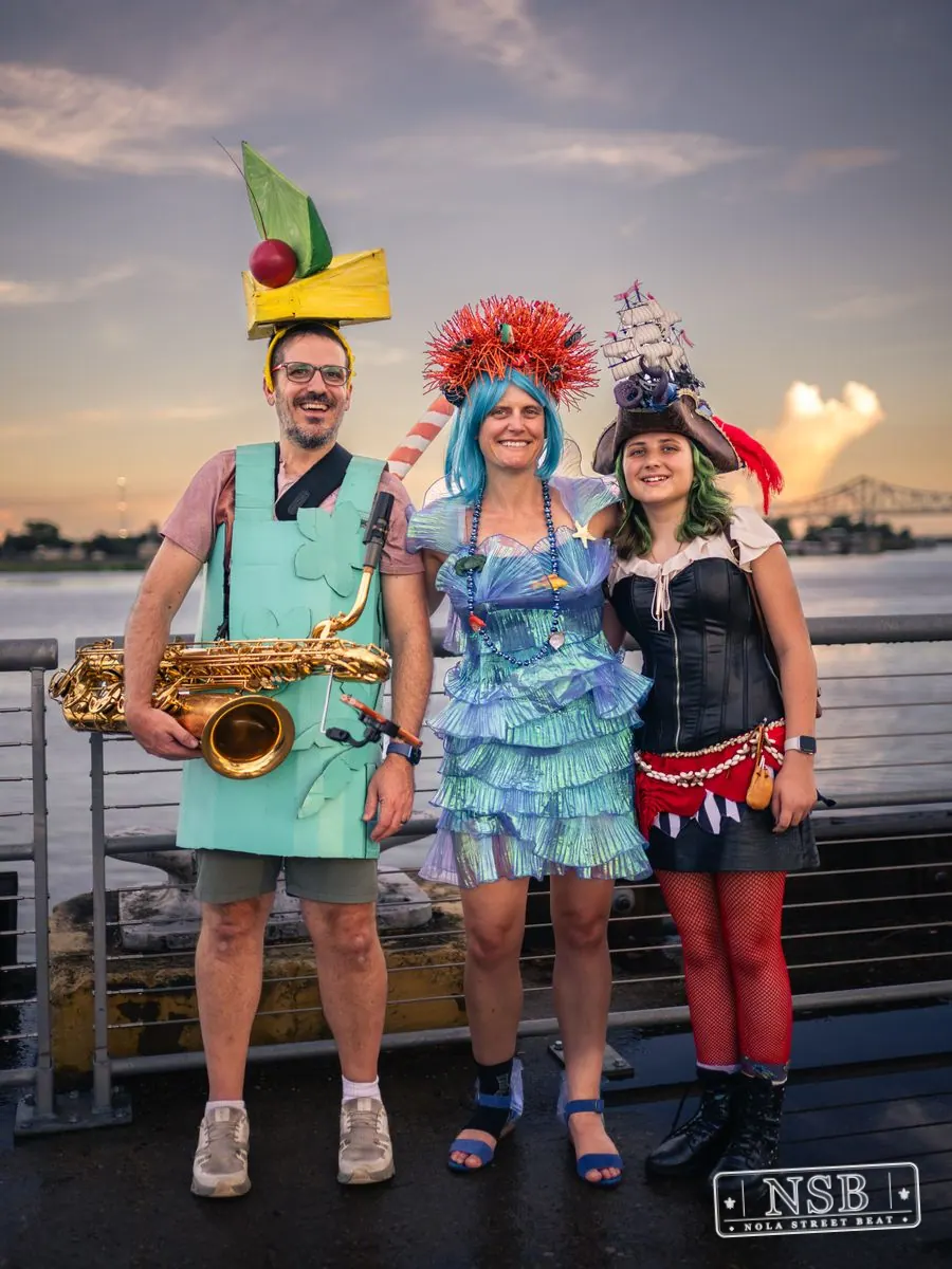 Three costumed parade musicians posing beside the river at the 2026 New Orleans Mermaid Parade
