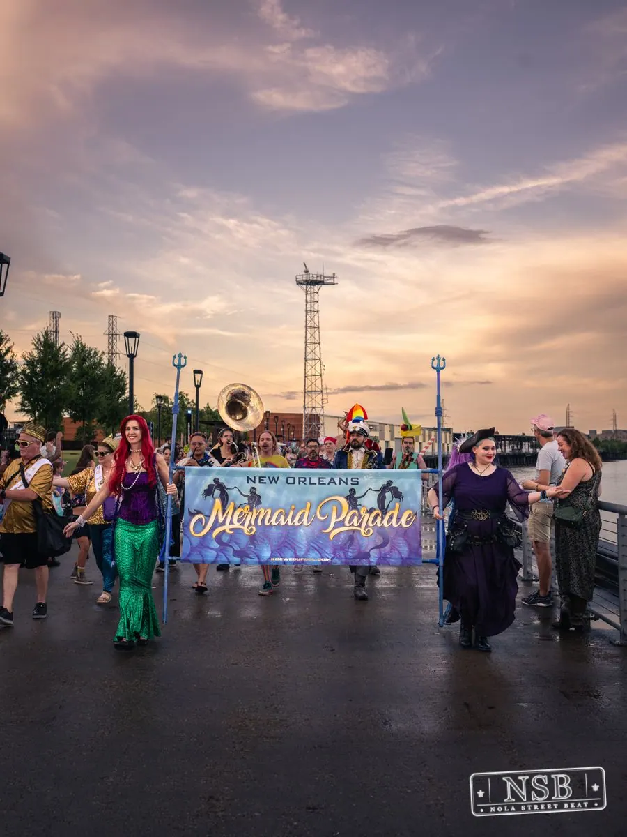 The Mermaid Parade gathering behind its banner on the riverfront promenade at the 2026 New Orleans Mermaid Parade