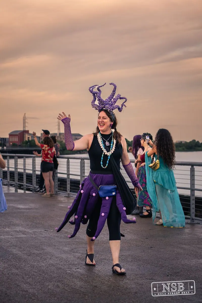 A participant in a purple tentacle-inspired headdress waving by the river at the 2026 New Orleans Mermaid Parade