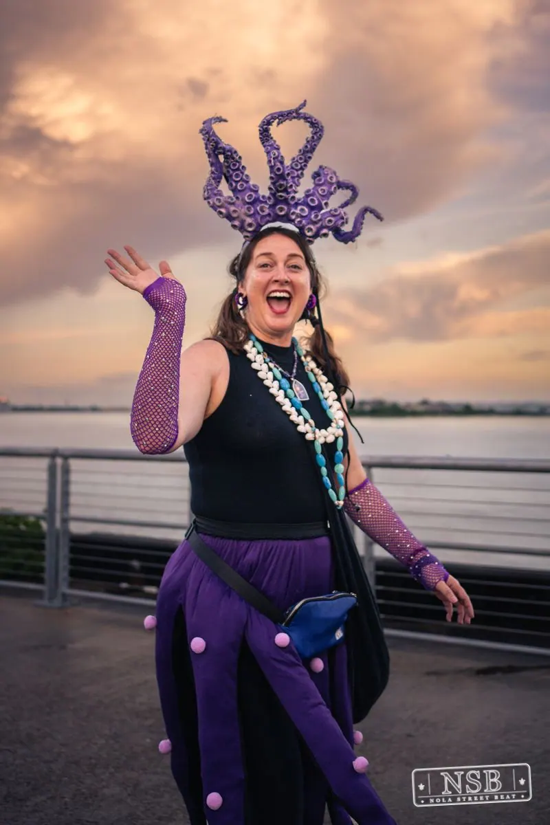 A participant in a purple tentacle-inspired headdress waving by the river at the 2026 New Orleans Mermaid Parade