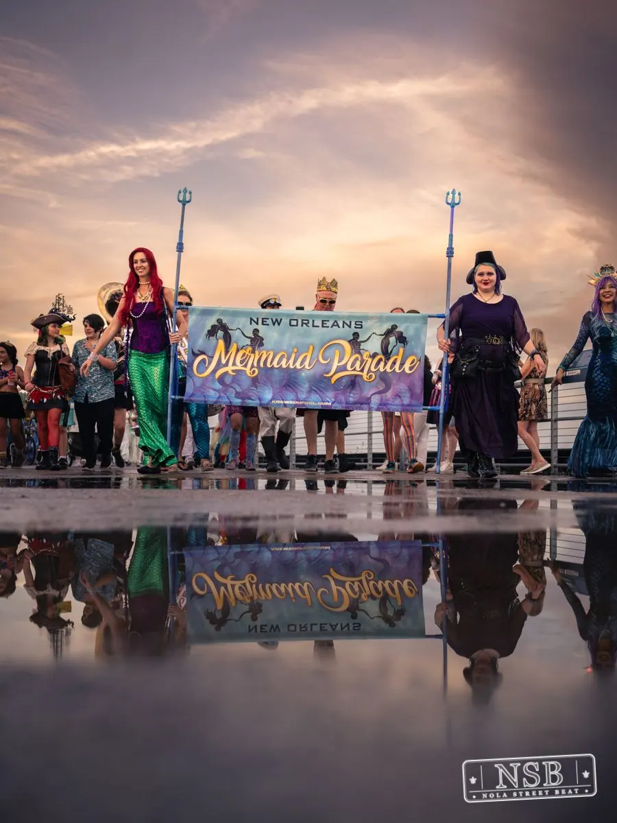 The Mermaid Parade banner and marchers reflected in rainwater on the promenade at the 2026 New Orleans Mermaid Parade