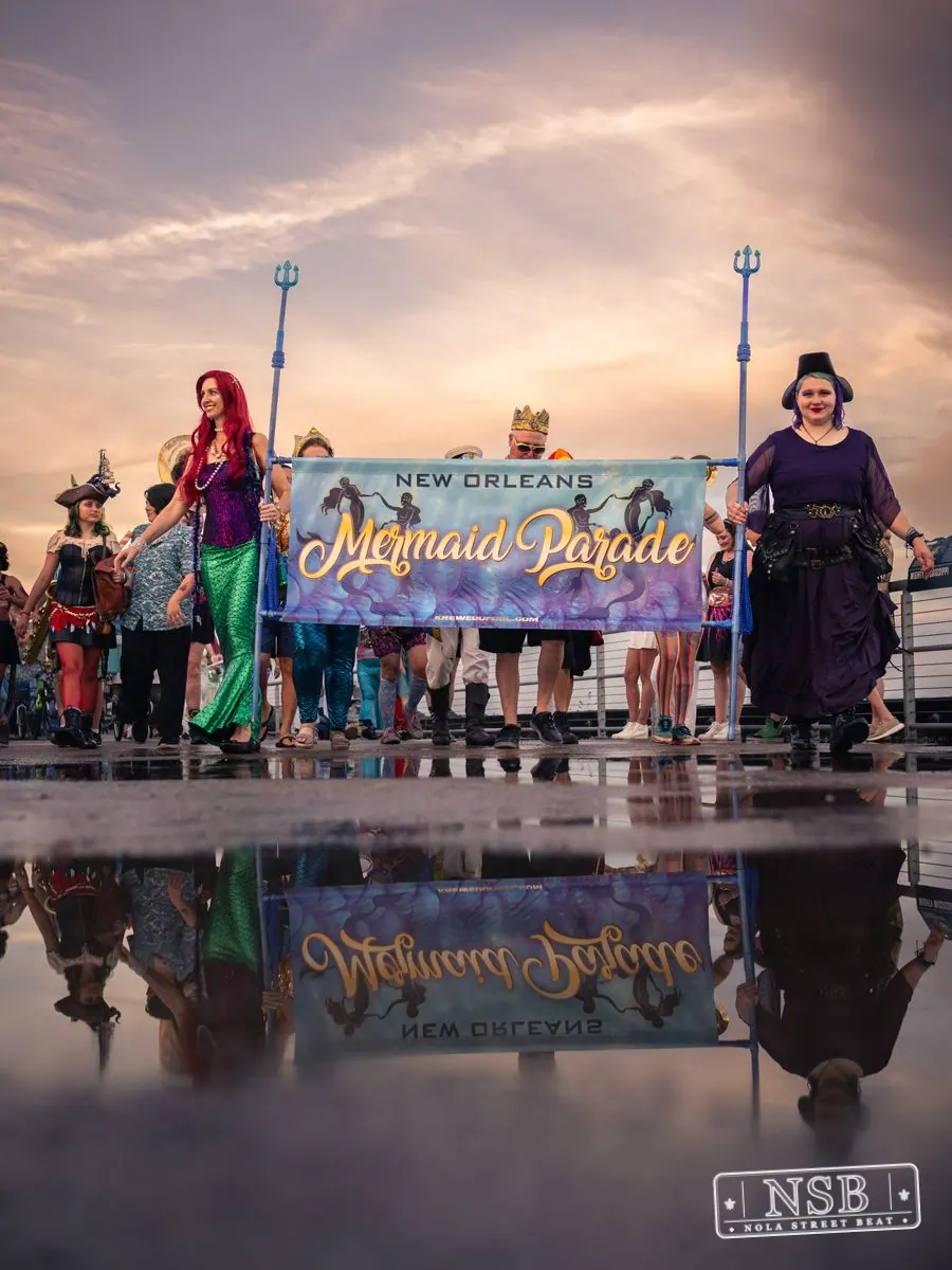 The Mermaid Parade banner and marchers reflected in rainwater on the promenade at the 2026 New Orleans Mermaid Parade