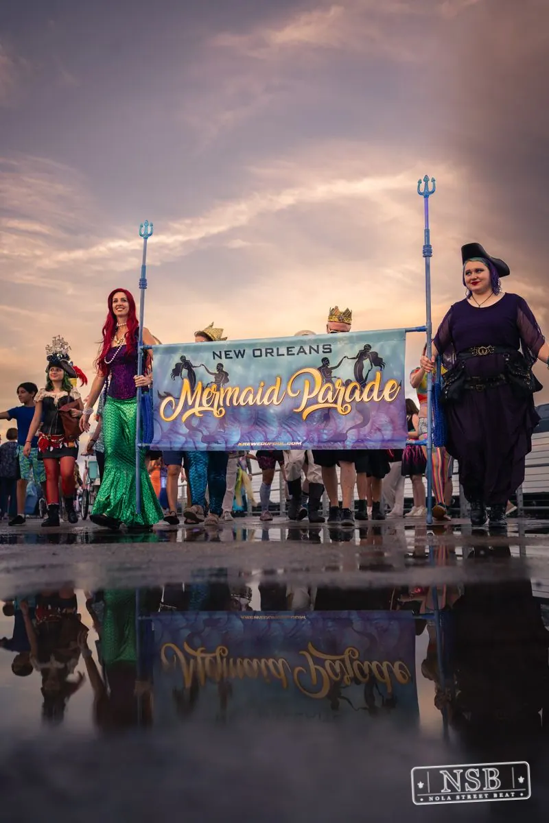The Mermaid Parade banner and marchers reflected in rainwater on the promenade at the 2026 New Orleans Mermaid Parade