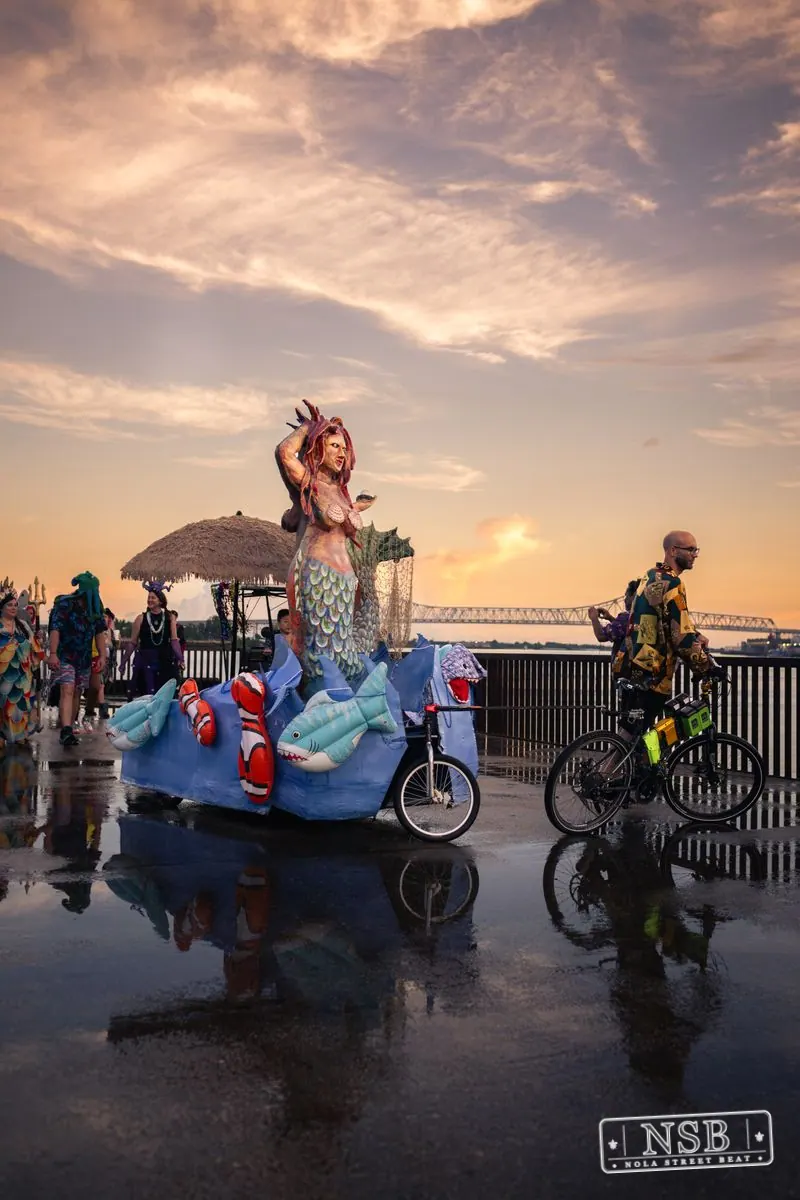 Large sea-themed figures and parade participants reflected on the wet promenade at the 2026 New Orleans Mermaid Parade