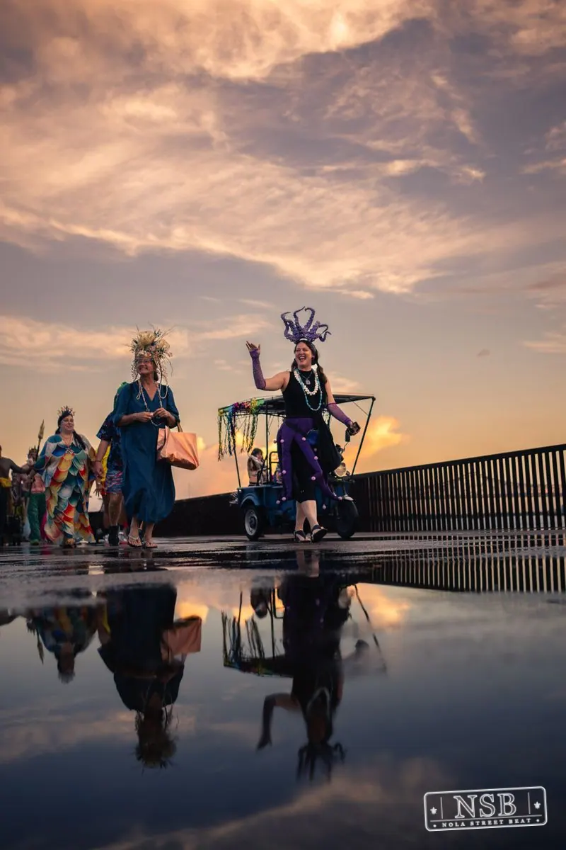 Large sea-themed figures and parade participants reflected on the wet promenade at the 2026 New Orleans Mermaid Parade