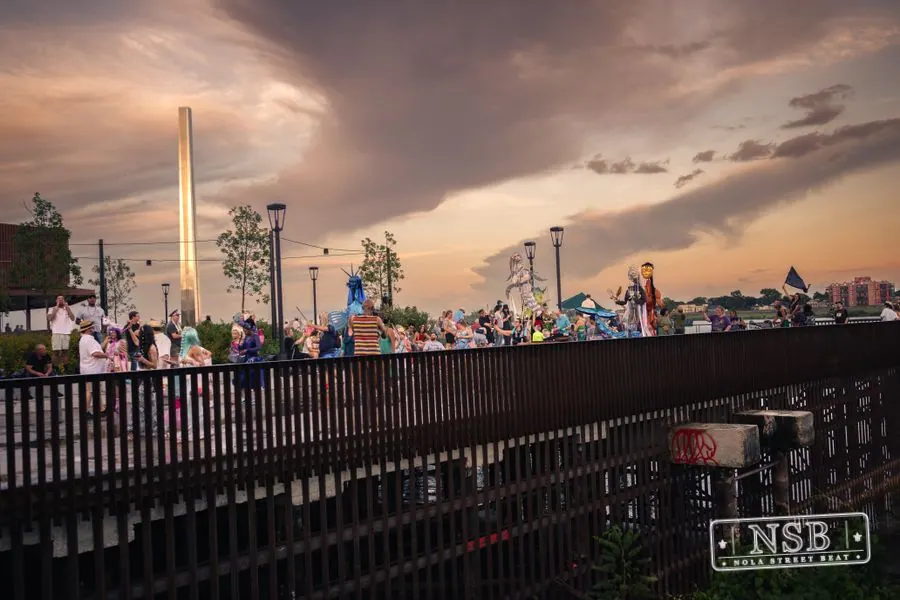 The parade crossing a riverfront walkway beneath a colorful evening sky at the 2026 New Orleans Mermaid Parade