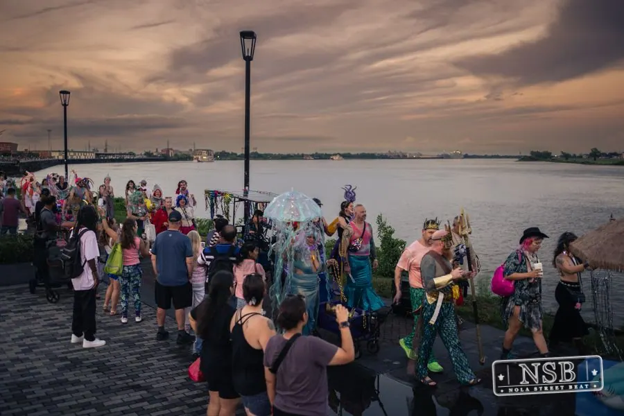 Costumed marchers filling the riverfront promenade as the sun sets at the 2026 New Orleans Mermaid Parade
