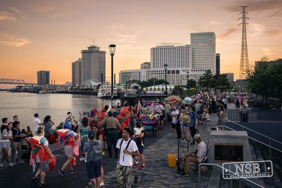 Costumed marchers filling the riverfront promenade as the sun sets at the 2026 New Orleans Mermaid Parade