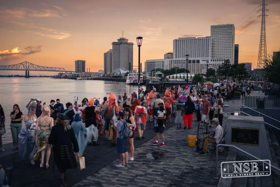 Costumed marchers filling the riverfront promenade as the sun sets at the 2026 New Orleans Mermaid Parade