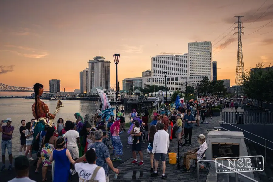 Costumed marchers filling the riverfront promenade as the sun sets at the 2026 New Orleans Mermaid Parade
