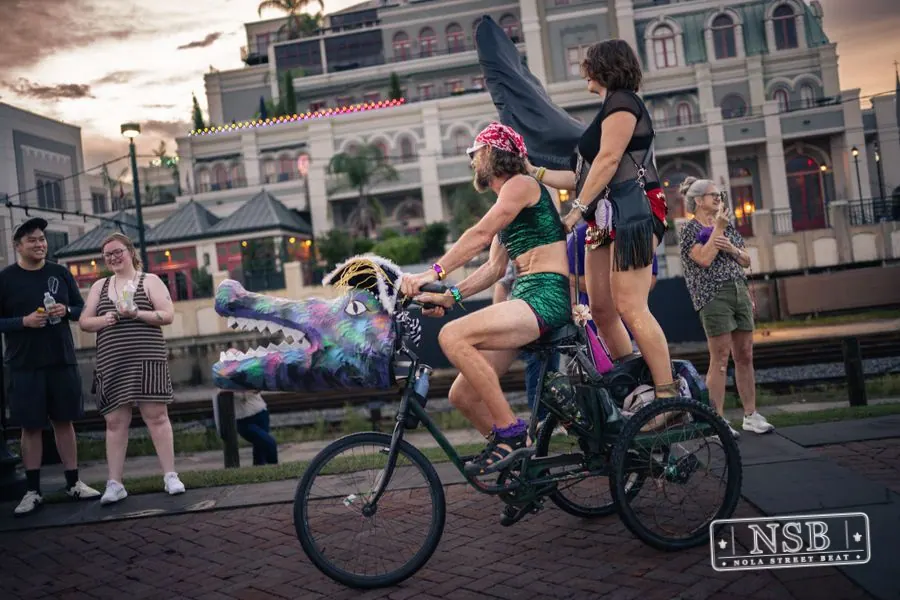 Decorated bicycles and a parade cart moving past riverfront buildings at the 2026 New Orleans Mermaid Parade