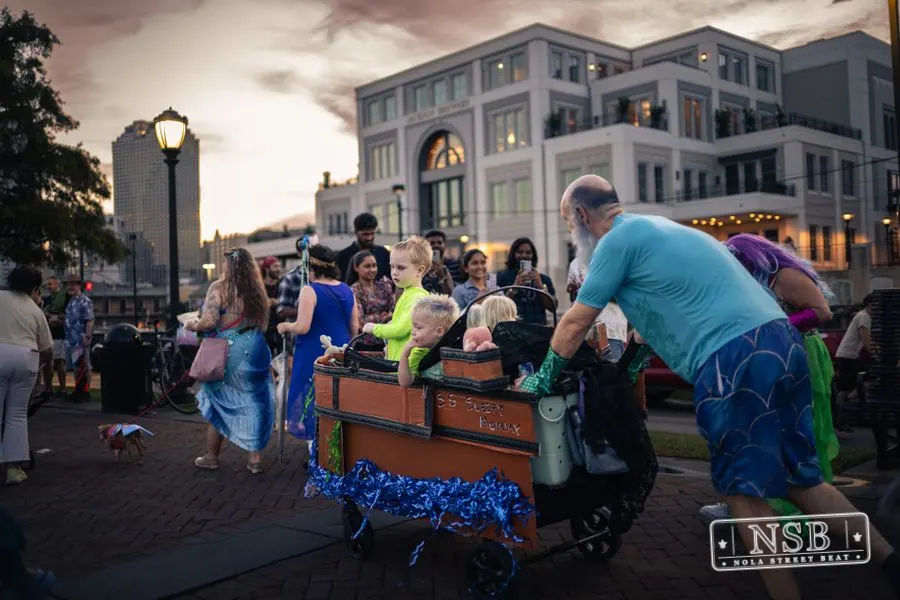 Decorated bicycles and a parade cart moving past riverfront buildings at the 2026 New Orleans Mermaid Parade