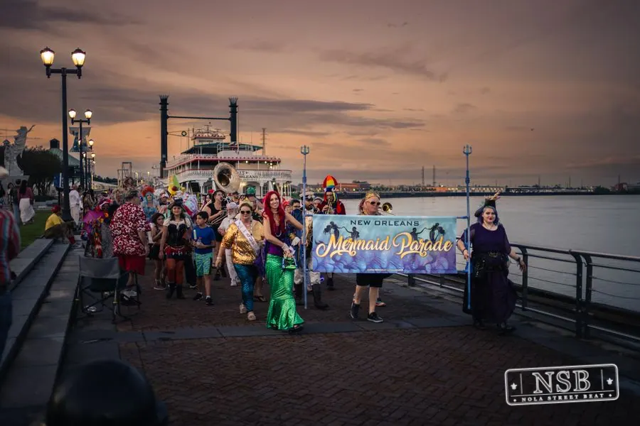 The Mermaid Parade banner and costumed marchers beside the river at sunset at the 2026 New Orleans Mermaid Parade