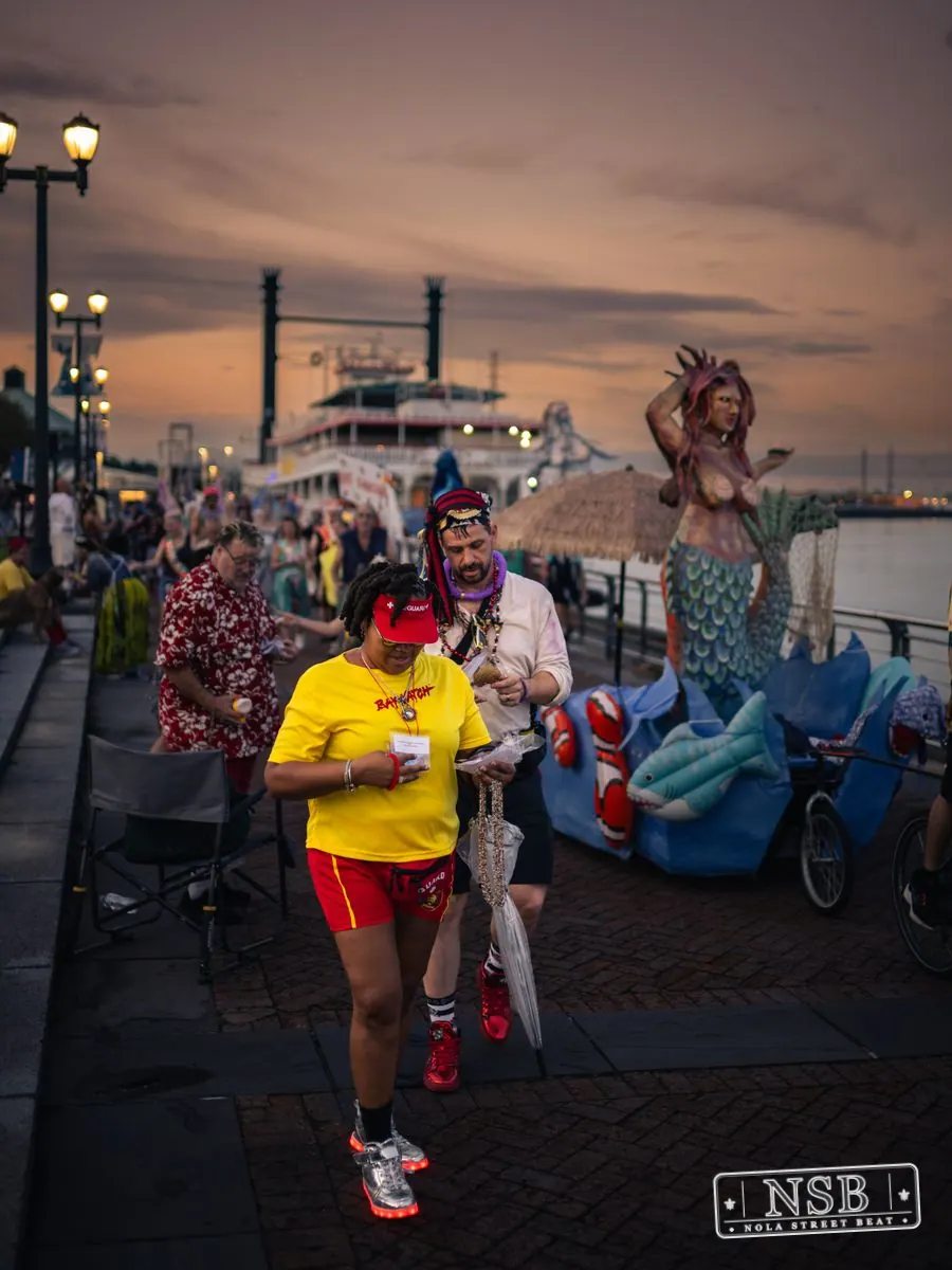 The Mermaid Parade banner and costumed marchers beside the river at sunset at the 2026 New Orleans Mermaid Parade