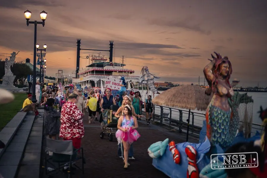 The Mermaid Parade banner and costumed marchers beside the river at sunset at the 2026 New Orleans Mermaid Parade