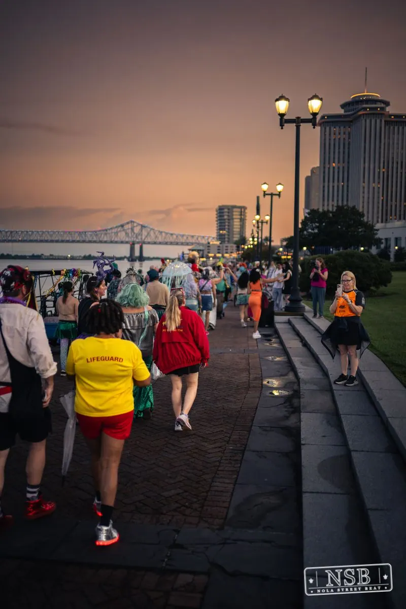 The Mermaid Parade banner and costumed marchers beside the river at sunset at the 2026 New Orleans Mermaid Parade