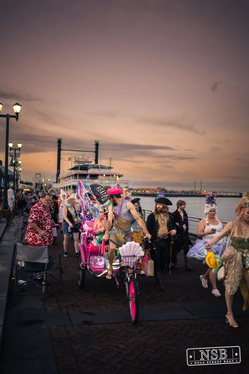 The Mermaid Parade banner and costumed marchers beside the river at sunset at the 2026 New Orleans Mermaid Parade