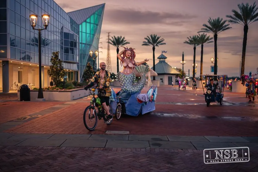 A decorated bicycle and mermaid figure passing the palm-lined Aquarium plaza at the 2026 New Orleans Mermaid Parade