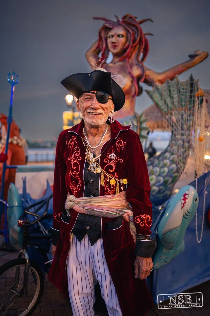 A pirate-costumed participant in front of a large mermaid figure at the 2026 New Orleans Mermaid Parade