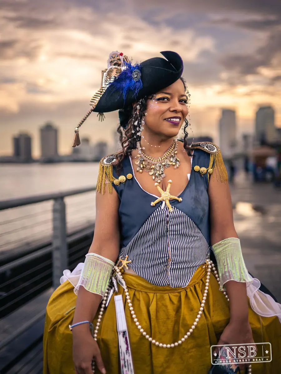 A pirate-inspired Mermaid Parade participant posing beside the Mississippi River with the New Orleans skyline behind her