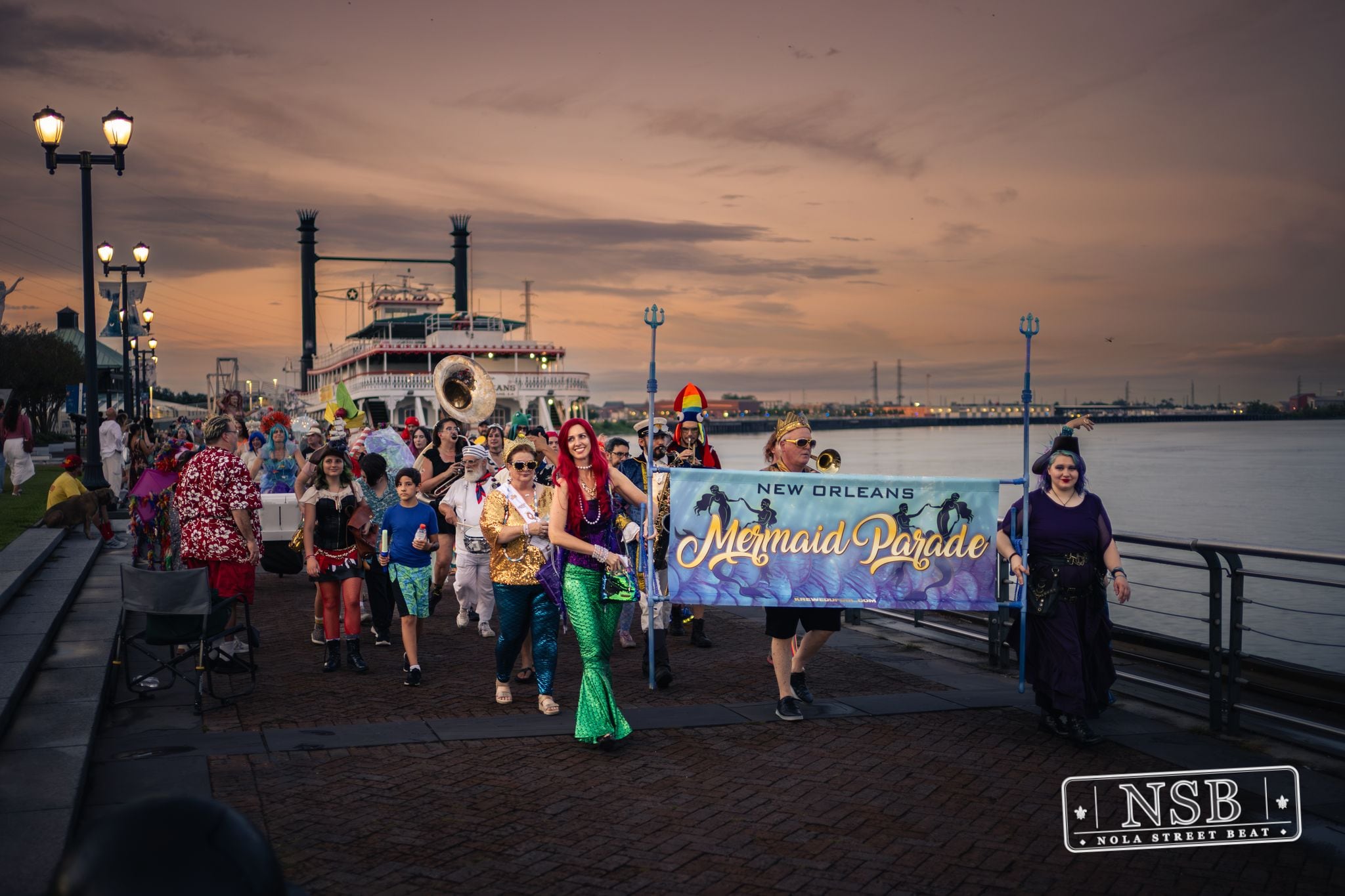 The Mermaid Parade banner and costumed marchers beside the river at sunset at the 2026 New Orleans Mermaid Parade
